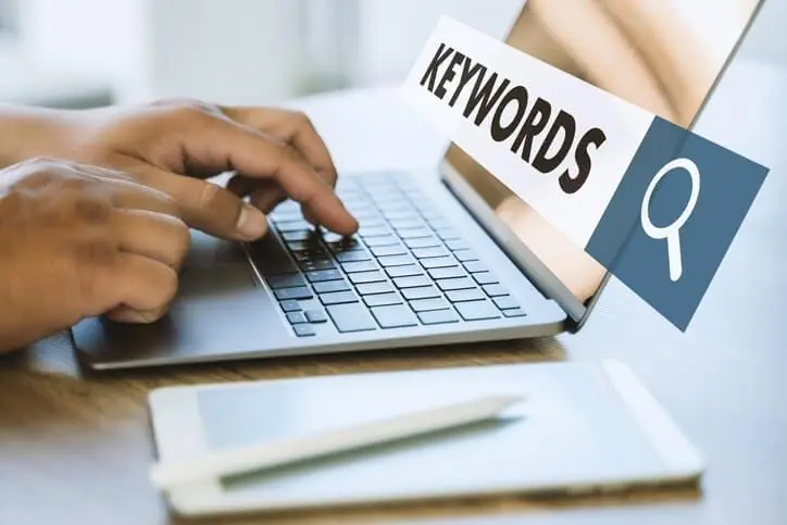 Pessoa digita em notebook com etiqueta Keywords e ícone de lupa, simbolizando a pesquisa de palavras-chave