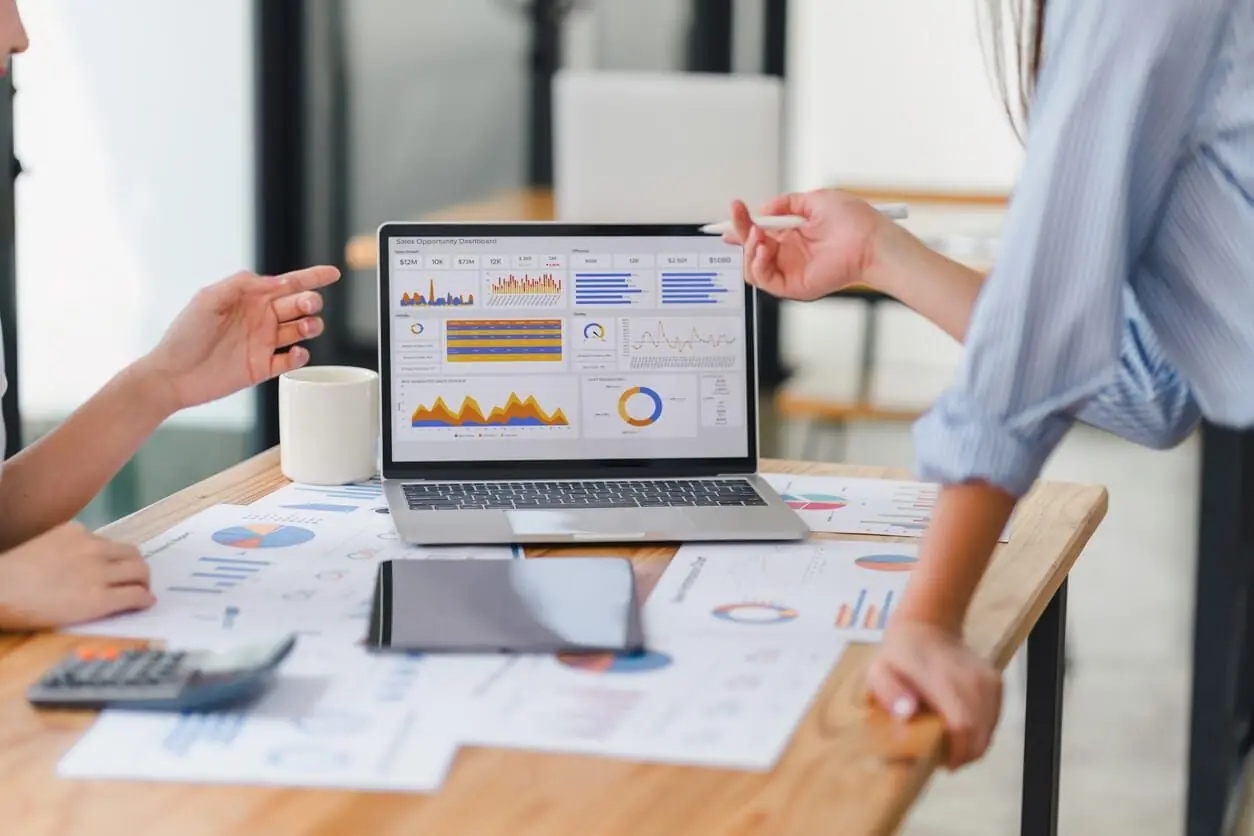 Duas pessoas analisam gráficos de um dashboard no notebook e relatórios impressos sobre a mesa
