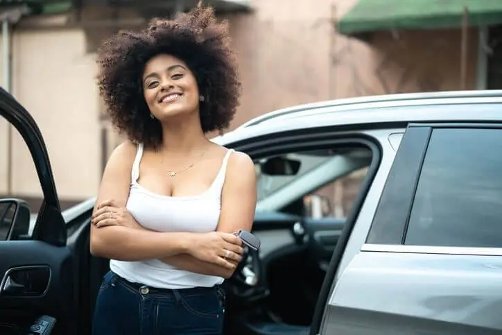 Mulher sorridente de braços cruzados segura a chave ao lado de carro prata com a porta aberta