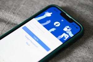 Smartphone exibindo a tela de login do Facebook, com plano de fundo em azul e ícones de interação (curtir, compartilhar), representando acesso a uma rede social.