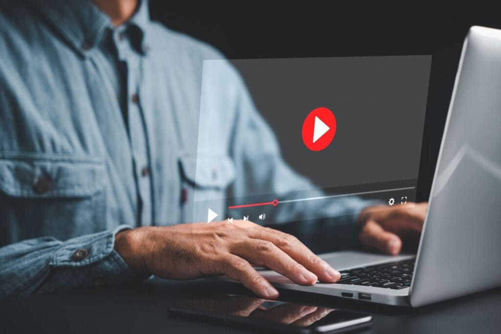 Homem vestindo camisa de mangas longas utilizando um notebook, com um ícone de reprodução de vídeo destacado na tela, sugerindo a criação ou a exibição de conteúdo multimídia.