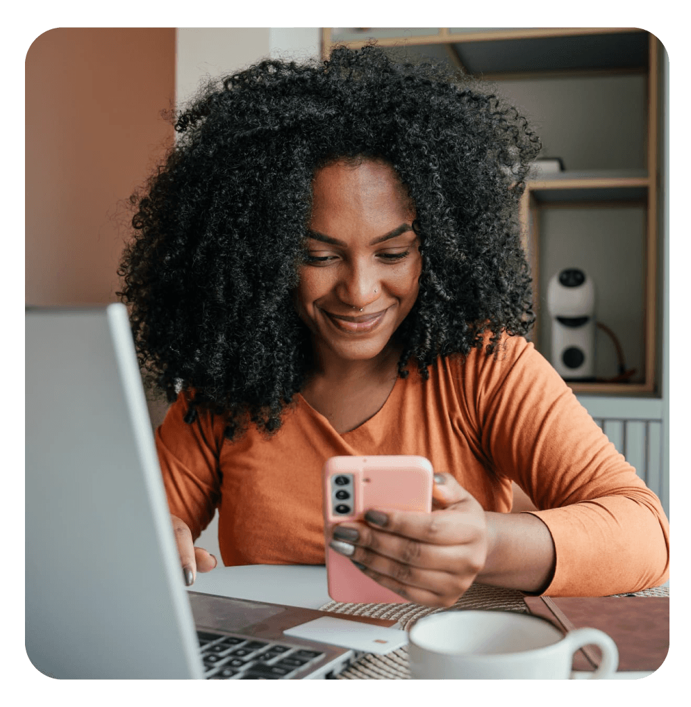 Mulher negra sorrindo enquanto usa um smartphone ao lado de um notebook, sugerindo trabalho remoto ou comunicação online.