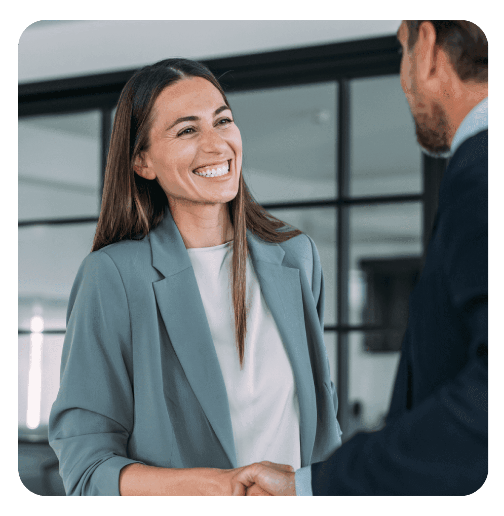 Mulher sorridente, usando blazer, cumprimentando um homem com um aperto de mãos em um ambiente profissional, simbolizando acordo ou parceria de negócios.