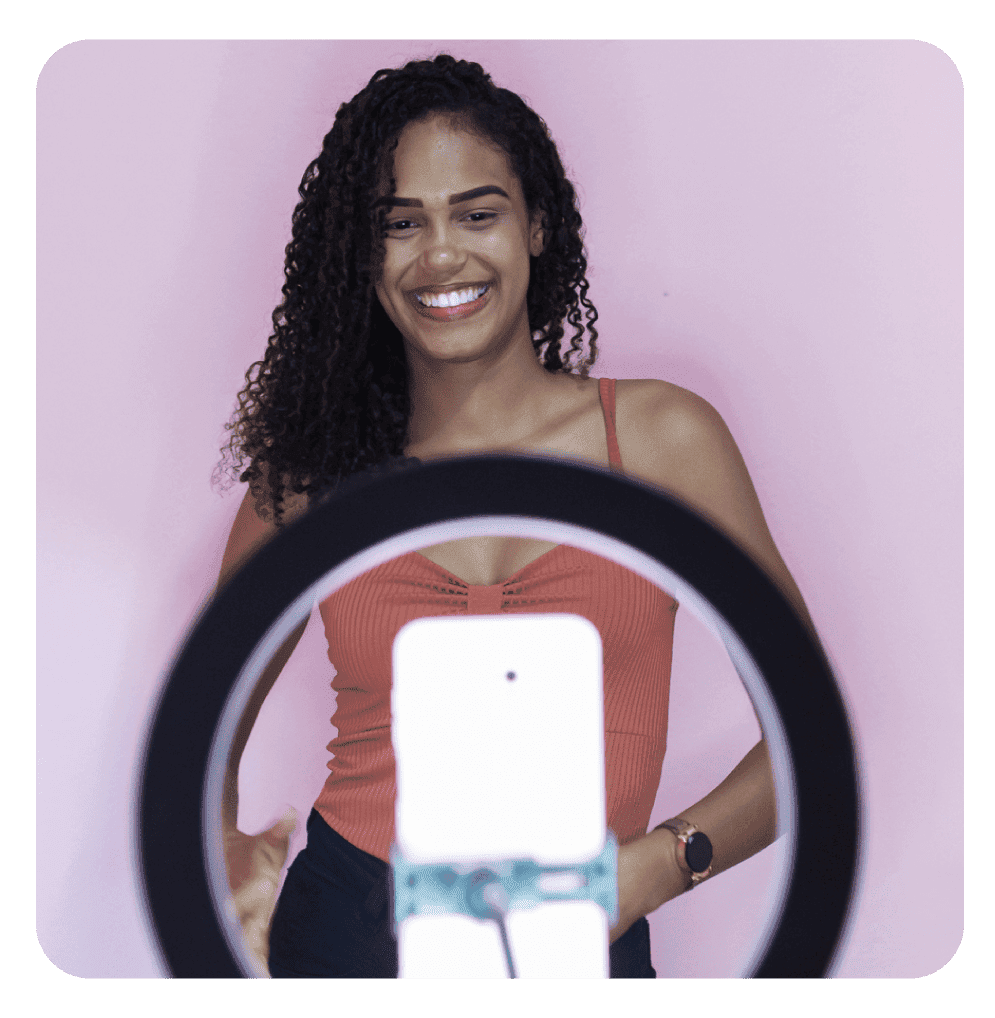 Mulher sorrindo em frente a um smartphone montado em um ring light, contra um fundo rosa, indicando gravação de conteúdo ou vídeo para redes sociais.